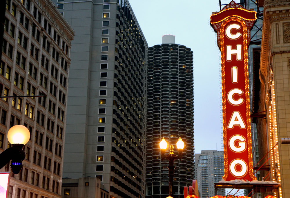 A large vertical Chicago Theatre sign glows in red and yellow lights amid tall downtown buildings at dusk, with street lamps and skyscrapers visible in the background.