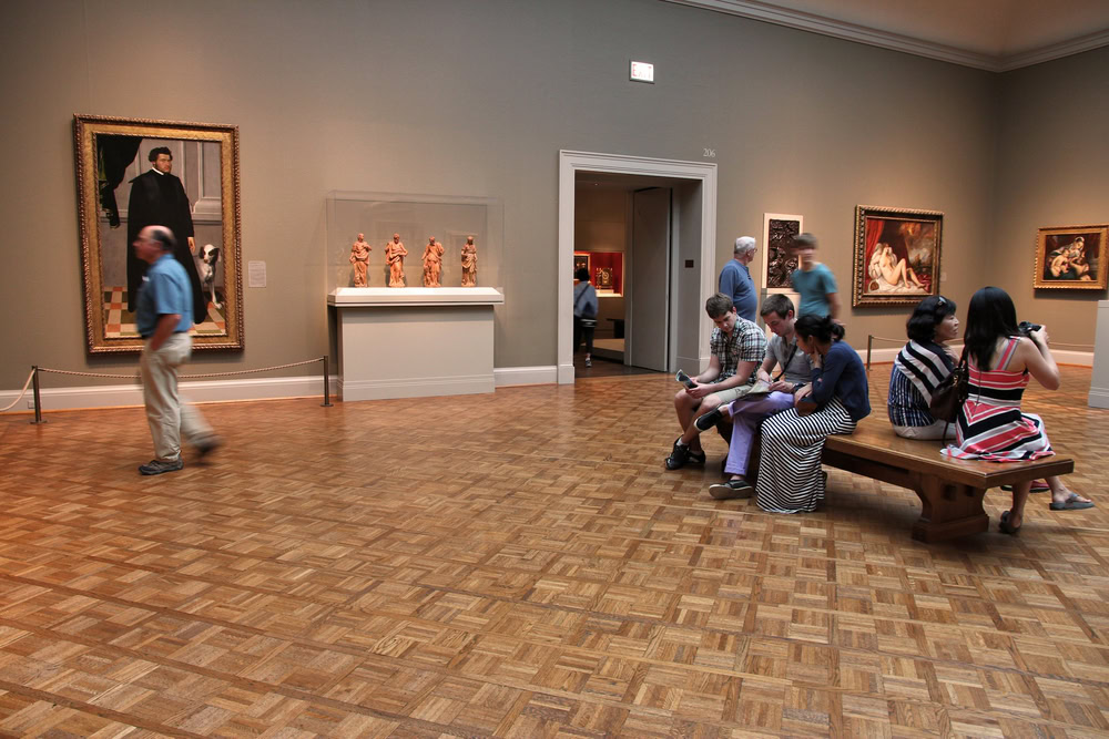 A spacious art gallery with several people observing paintings and sculptures; some are sitting on a wooden bench, while others are walking or standing and viewing the artwork displayed on the walls and in a glass case.