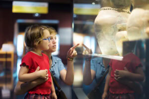 An adult holding a young child in front of a glass display case at a museum, both looking closely at ancient pottery while the adult points at the exhibit.