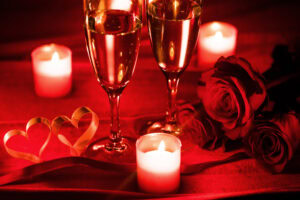 Two glasses of champagne, three lit candles, two red roses, and two ribbon hearts sit on a red surface, creating a romantic atmosphere with warm lighting.