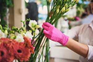 A person wearing bright pink gloves arranges tall white flowers in a vase, with other flowers visible in the foreground. The setting appears to be a flower shop or floral workspace.