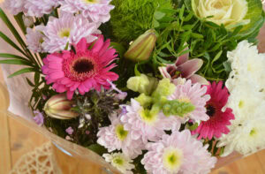 A bouquet of assorted flowers including pink gerbera daisies, white chrysanthemums, green foliage, and flower buds, arranged closely together on a wooden surface.