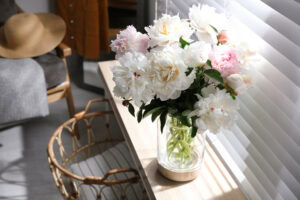 A glass vase filled with white and light pink peonies sits on a wooden table near a window with closed blinds. A wicker basket and a chair with a hat and blanket are nearby. Sunlight filters through the blinds.