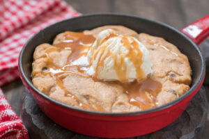 A warm chocolate chip cookie baked in a red skillet, topped with a scoop of vanilla ice cream and drizzled with caramel sauce, sits on a wooden surface next to a red and white checkered cloth.