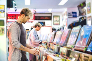 Two people browse through vinyl records in a brightly lit record store, surrounded by colorful album covers and posters on the walls.