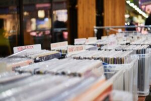 Plastic bins filled with rows of vinyl records, each separated by labeled dividers such as “KRAUTROCK” and “UK 60’S/70’S ROCK,” displayed in a brightly lit record store.