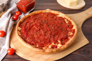 A Chicago-style deep dish pizza topped with chunky tomato sauce sits on a wooden pizza peel, with a glass dish of extra sauce, a block of cheese, and cherry tomatoes nearby on a wooden table.