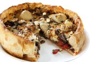 A deep dish pizza with a thick, golden crust, topped with artichoke hearts, mushrooms, cheese, and vegetables, sits on a white plate. A large slice has been cut and slightly removed from the whole pizza.