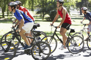 Four people wearing helmets ride bicycles together on a sunny day in a park, with trees and greenery in the background. Two are in the foreground, one in a blue shirt and one in a red shirt.