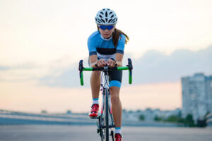 A cyclist in a blue and black outfit, helmet, and sunglasses rides a road bike on an urban street at dusk, with blurred buildings and sky in the background.
