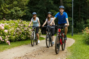 Three people wearing helmets ride bicycles on a dirt path through a park with trees and pink flowers, smiling and enjoying the outdoors.