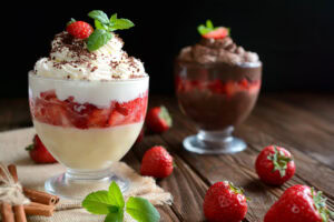 Two glass cups filled with layered desserts; the front one has vanilla pudding, strawberries, whipped cream, chocolate shavings, and a mint garnish, while the back one features chocolate mousse with strawberries. Fresh strawberries are scattered around.