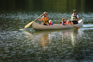 Four people wearing life jackets paddle a canoe on a calm lake, surrounded by green reflections. Two adults are paddling at the ends, and two children sit in the middle, enjoying the ride.