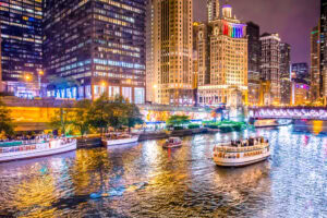 Downtown cityscape at night with tall buildings lit up, colorful lights reflecting on a river, and several boats cruising along the water near a bridge.