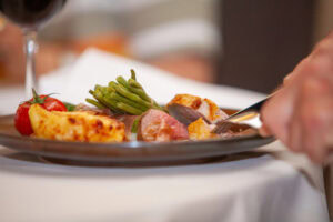 A close-up of a person cutting into a plated meal with a fork and knife, featuring roasted meat, green beans, grilled tomatoes, and a cheesy potato side, on a white tablecloth.