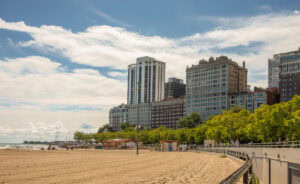 Tall buildings and lush trees line a sandy beach with a partly cloudy sky above. A few people are seen on the beach, and a boardwalk runs along the right side of the image.