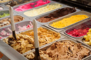 A display case filled with various colorful gelato flavors, including chocolate, vanilla, strawberry, and pistachio, with metal scoops placed in some of the tubs.