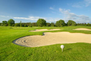 A golf ball on a tee sits on a lush green course near a large sand bunker under a blue sky with scattered clouds and surrounding trees in the background.