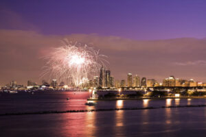 Fireworks light up the sky over a city skyline at dusk, with tall buildings illuminated and reflected in the water; a lighthouse stands near the waterfront in the foreground.