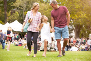 A smiling family of three walks hand in hand on grass at an outdoor event, with tents and a crowd in the background. The woman carries a tote bag with baguettes, and the young girl looks up at her parents.