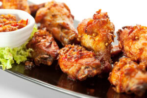Spicy, glazed chicken wings are arranged on a black plate with a small bowl of chunky dipping sauce and a garnish of fresh lettuce on the side.