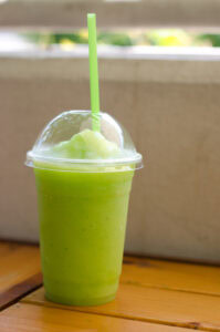A bright green smoothie or slush in a clear plastic cup with a dome lid and green straw, placed on a wooden table with a blurred outdoor background.