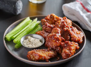 A plate of saucy buffalo chicken wings is served with celery sticks and a small dish of creamy dipping sauce, garnished with black pepper, on a dark table next to a folded white towel.