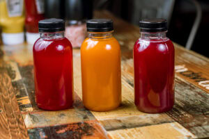 Three small plastic bottles with black caps, filled with red and orange liquids, are placed on a rustic wooden table. The background is blurred with condiment bottles visible.