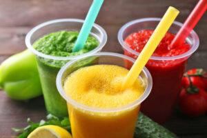 Three plastic cups filled with colorful smoothies—green, yellow, and red—each with a matching straw. Fresh fruits and vegetables, including a lemon, tomato, and cucumber, are placed beside the drinks on a wooden surface.