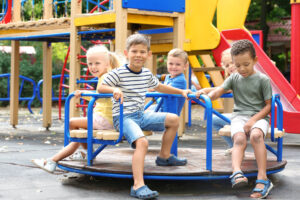 Five young children sit and smile on a blue and yellow playground merry-go-round, surrounded by outdoor play structures on a sunny day. They appear to be enjoying their time together.