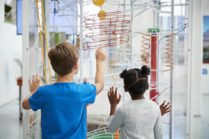 Two children, a boy in a blue shirt and a girl with pigtails, view and interact with a large kinetic ball machine exhibit featuring winding tracks and colorful elements in a science museum.