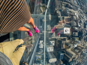 Two people stand on a glass observation deck high above a city, looking down at skyscrapers and streets far below. One person wears a striped dress and pink shoes; the other wears pants and gray shoes.