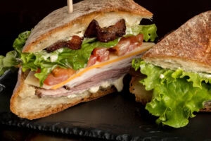 A close-up of a sandwich with layers of lettuce, tomato, cheese, turkey, ham, bacon, and mayonnaise on rustic bread, served on a dark slate surface.