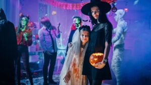 A woman dressed as a witch and a girl in a bloody white dress pose with a carved pumpkin at a Halloween party. Other costumed people, including a mummy, are dancing in the background amid festive decorations.