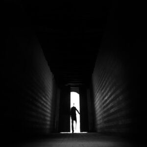 A person silhouetted in darkness walks toward a bright open doorway at the end of a narrow, shadowy corridor with striped walls.