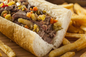 A close-up of an Italian beef sandwich topped with chopped giardiniera vegetables served in a white hoagie roll, with a side of golden French fries on a wooden surface.