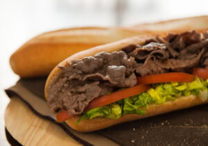 A close-up of a sandwich filled with sliced roast beef, shredded lettuce, and tomato slices on a baguette, resting on a cloth napkin with another baguette in the background.
