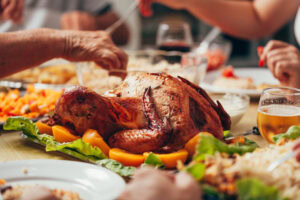 A roasted turkey garnished with vegetables sits on a table surrounded by people enjoying a meal together. Plates, drinks, and hands reaching for food are visible, suggesting a festive gathering or celebration.