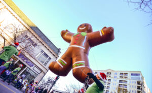 A large gingerbread man balloon floats above a street during a parade, held by people wearing green shirts and Santa hats, with buildings and spectators in the background.