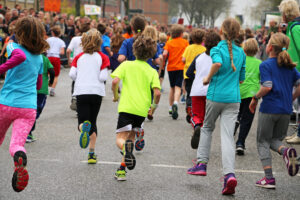 A group of children in colorful clothing are running together on a street during a race or event, with a crowd of spectators watching in the background.