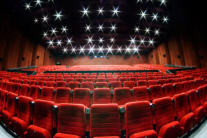 A wide-angle view of an empty theater with rows of red seats facing a stage. The ceiling features bright, star-shaped lights, and the walls are dark brown.