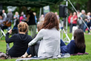 Three people sit on the grass with their backs to the camera at an outdoor event. A crowd and stage are visible in the background, with trees and a canopy overhead. The atmosphere appears relaxed and social.