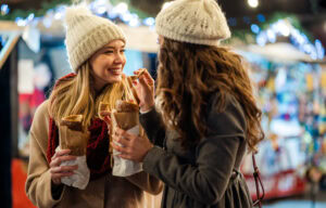 Two women in winter coats and knit hats smile and enjoy food wrapped in paper at an outdoor market decorated with string lights and festive decorations.