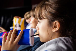 A young girl in a movie theater drinks from a large blue cup with a straw, while other children in the background hold popcorn.
