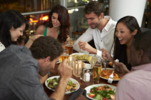 A group of five friends sits around a table at a restaurant, laughing and enjoying plates of pasta and salad, with drinks and candles on the table.