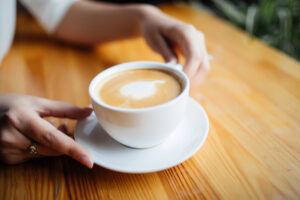 A person holds a white cup of coffee with heart-shaped latte art on a wooden table, with one hand on the cup and the other resting nearby.