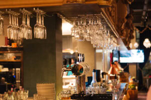 A bar interior with rows of hanging wine glasses above a counter, beer taps, cups, and various bar equipment. Warm lighting creates a cozy atmosphere, and a TV is visible in the background.