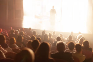 An audience sits in a dark theater watching a brightly lit stage, where a single performer is visible in silhouette against a glowing background.