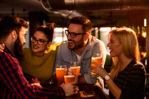 Four friends sit at a bar table, smiling and laughing while holding pints of beer. The group appears to be enjoying each other’s company in a warm, dimly lit setting.
