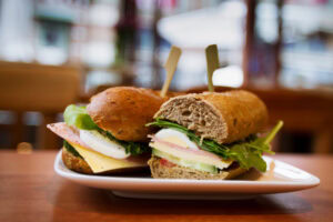 A whole grain sandwich cut in half on a white plate, filled with sliced boiled egg, cheese, lettuce, and ham, set on a wooden table with a blurred café background.
